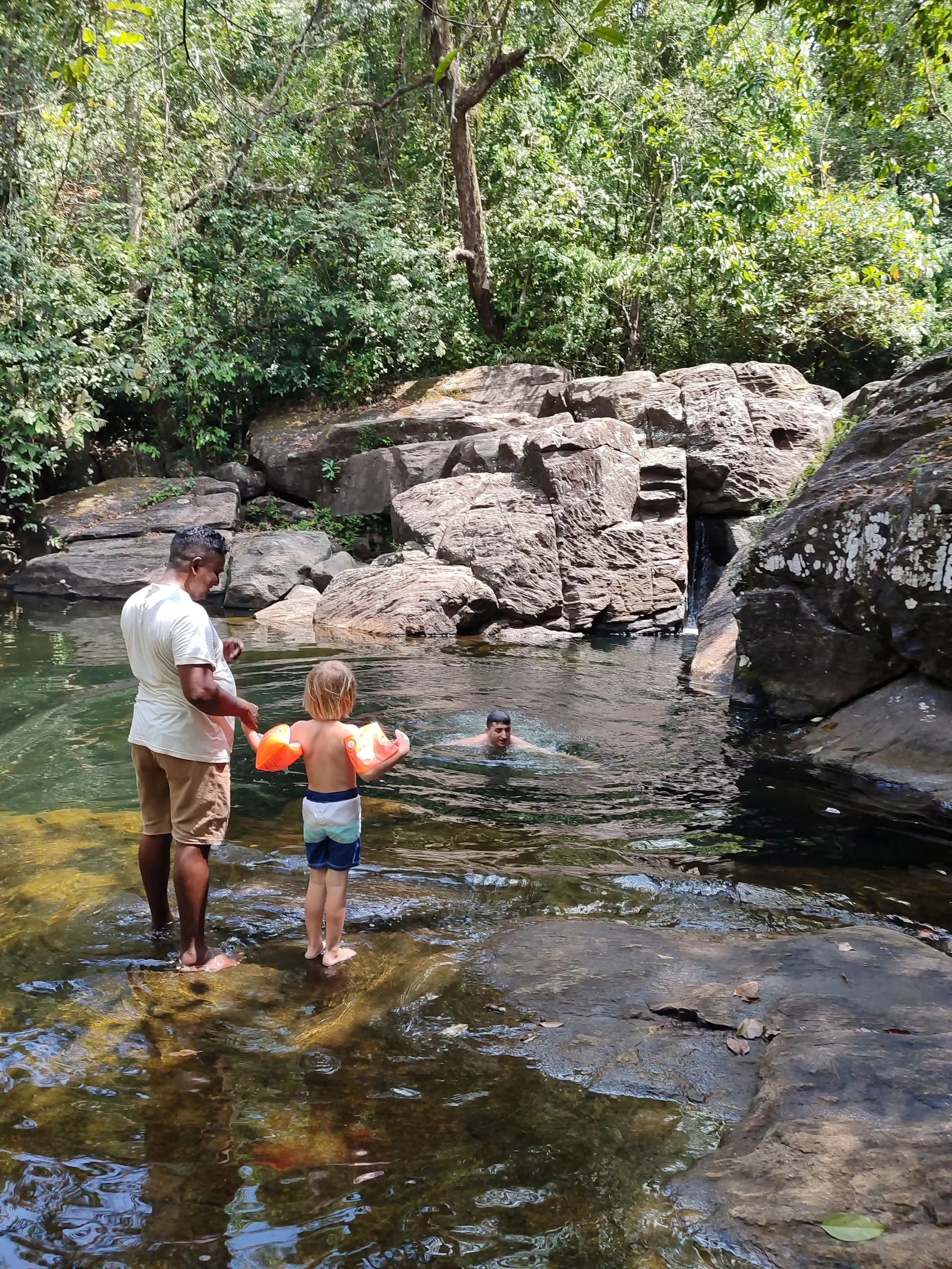 Ein Guide hält einen kleinen Jungen an der Hand am Rand eines Dschungelbeckens, dahinter schwimmt ein Mann