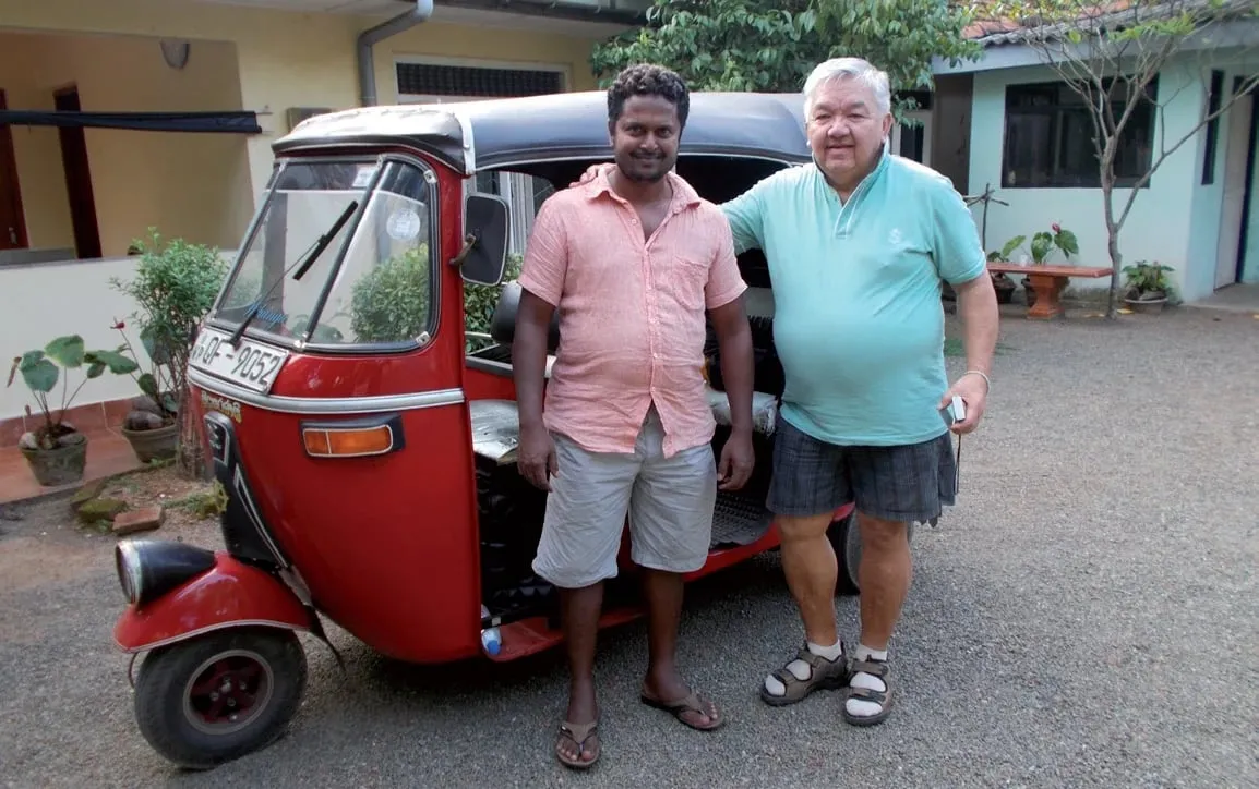 Tony Achmat and Kumar standing side by side in front of a red tuk-tuk at the family house in Aluthgama